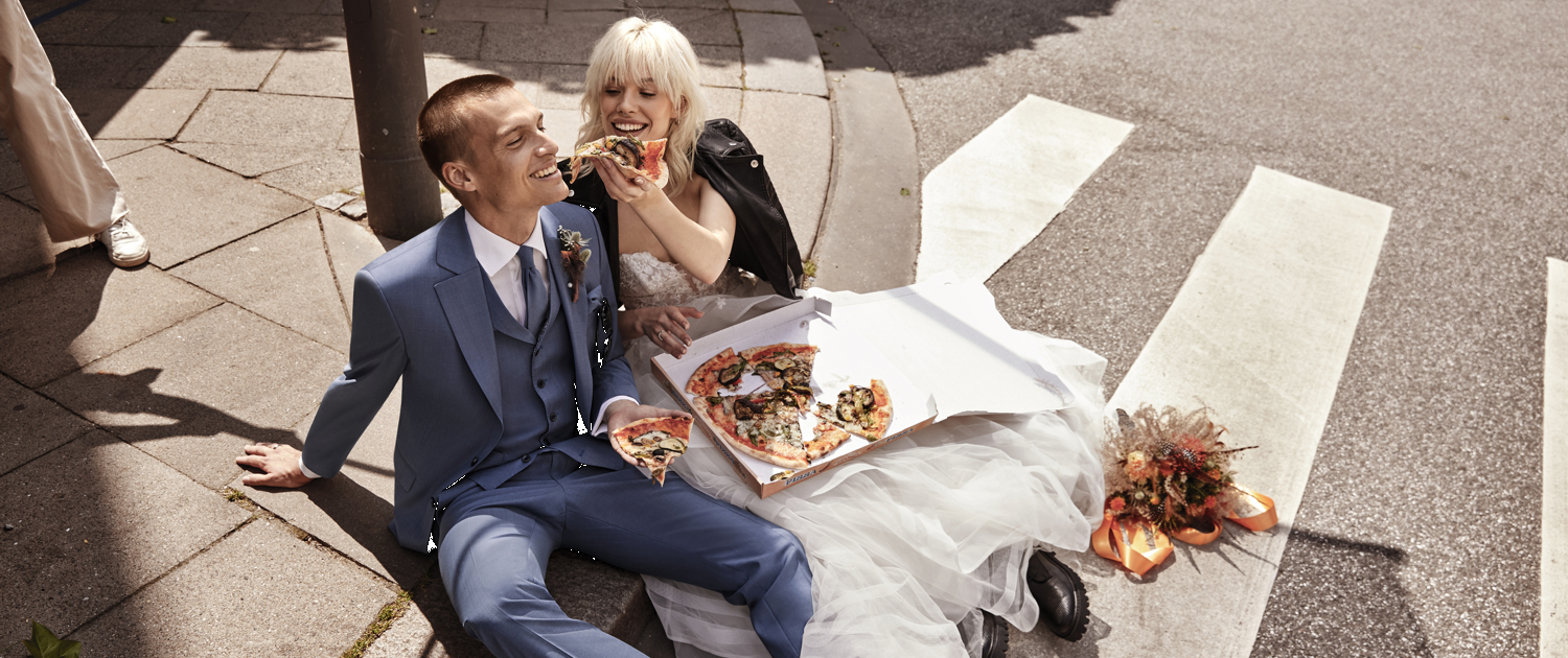 Hochzeitspaar auf dem Asphalt sitzend füttert sich mit Pizza